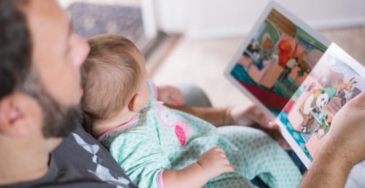 A baby sits on their father's lap while he reads from a picture book. The baby is wearing a light blue patterned onesie. The photo is taken from an angle looking over the father's shoulder from above.  