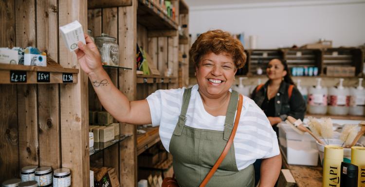 A smiling woman with brown skin and short hair reaches for a box on a shelf in a shop.  