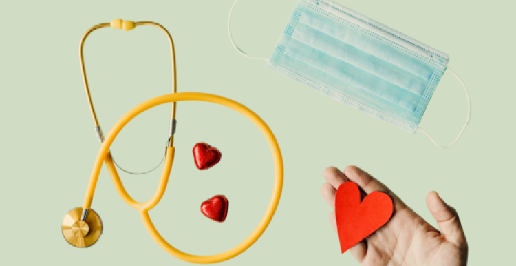 A hand holding a red paper heart, a stethoscope, two heart shaped chocolates in red foil, and a surgical mask spread across a light background. 