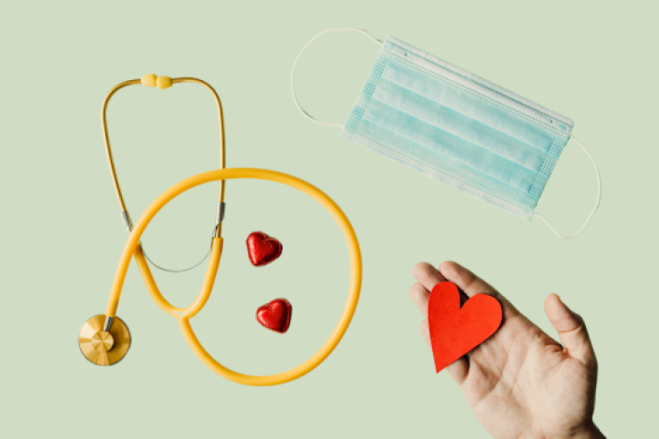 A hand holding a red paper heart, a stethoscope, two heart shaped chocolates in red foil, and a surgical mask spread across a light background. 